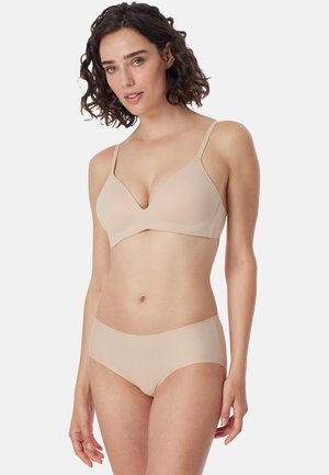 Donna con capelli corti e ricci che indossa un reggiseno senza cuciture color nude e biancheria intima coordinata su uno sfondo chiaro e uniforme.