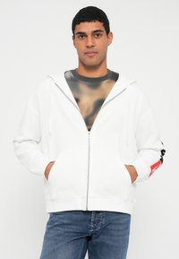 Balta kapuci jaka ar rāvējslēdzēju, priekšpusē kabata un kapucis ar auklu, piedurknēs melnas detaļas, valkāta virs rakstaina pelēka krekla.