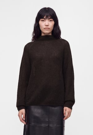 Kvinde med sort bølget hår iført en mørkebrunt langærmet sweater og en sort lædernederdel, stående mod en hvid baggrund.