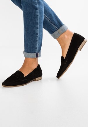 Sorte ruskinds slip-ons med en lav træhæl og en dekorativ rem. Sammen med oprullede blå denimjeans. Hvid baggrund.