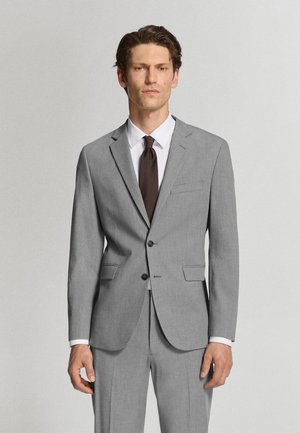 Hombre con un traje gris claro a medida, camisa blanca y corbata marrón oscuro, de pie frente a un fondo sencillo y claro.
