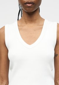 Άνω σώμα ατόμου με κοτσίδες, που φοράει λευκό αμάνικο τοπ με V-λαιμόκοψη και έχει μικρό τατουάζ στο εσωτερικό του αριστερού βραχίονα.