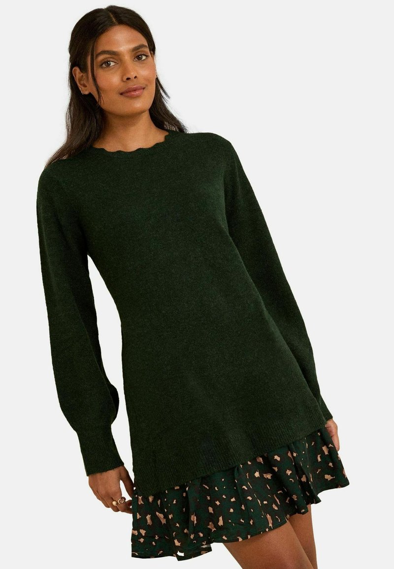 Temno zeleno pleteno sv sweater z dolgimi rokavi in valovitim ovratnikom, položen over črno krilo z bež abstraktnimi vzorci.