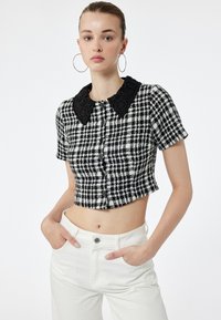 Zwart-witte geblokte cropped top met een zwarte geborduurde kraag, korte mouwen en een voorknoopsluiting, gecombineerd met witte broeken.