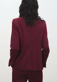 Burgundský přizpůsobený blazer s dlouhými rukávy, vypasovaným střihem a klopami. Hladká textura s jedním rozparkem na zadní straně a knoflíkovými manžetami.