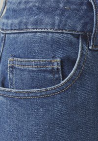 Jean en denim bleu avec une taille mi-haute, comprenant une petite poche arrière et des coutures jaunes contrastantes le long des coutures et des bords.