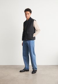 Schwarze steppbare Weste über einem beigen Langarmshirt, kombiniert mit blauen Jeans und schwarzen Schuhen. Schlichtes Design mit zwei Fronttaschen.