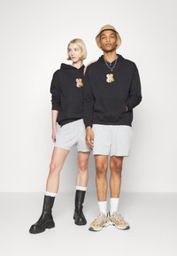 Black hoodies feature a cartoon character graphic on the chest. Paired with gray shorts, one model wears black boots; the other sports colorful sneakers.
