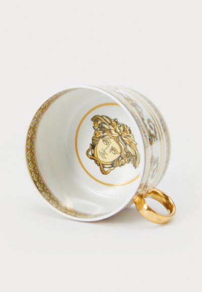 Λευκό κεραμικό φλιτζάνι με χρυσές λεπτομέρειες, που διαθέτει διακοσμητικό χρυσό σχέδιο και σχεδιασμένο πρόσωπο στο εσωτερικό. Η λαβή είναι χρυσή και στρογγυλεμένη.