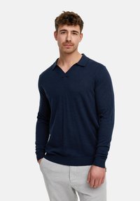 Dunkelblauer langärmliger Polo-Sweater mit V-Ausschnitt, gerippten Bündchen und Saum. Weicher Strickstoff mit kleinem Logo auf der Brust. Kombiniert mit hellen Hosen.