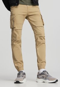 Beżowe spodnie cargo z elastycznymi mankietami, noszone z szarymi i białymi trampkami oraz białymi skarpetkami, na jednolitym jasnoszarym tle.