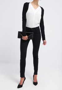 Frau in einem schwarz-weißen Outfit, die eine schwarze Clutch hält, schlanke schwarze Hosen und schwarze High Heels trägt und vor einem schlichten Hintergrund steht.