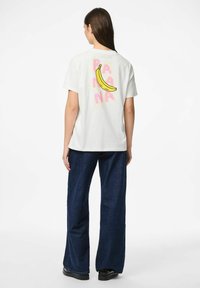 Valkoinen puuvillapaita, jossa on värikäs banaanigrafiikka ja sana "BANANA" vaaleanpunaisella, yhdistettynä sinisiin leveisiin farkkuihin.