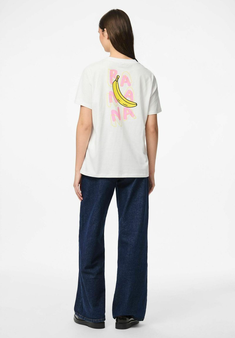 Valkoinen puuvillapaita, jossa on värikäs banaanigrafiikka ja sana "BANANA" vaaleanpunaisella, yhdistettynä sinisiin leveisiin farkkuihin.