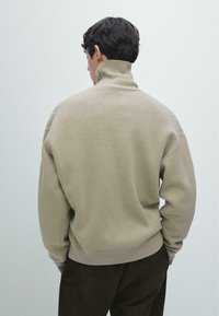 Beiger Pullover mit hohem Kragen aus weichem Strickstoff, mit lockerem Schnitt, überschnittenen Schultern und geripptem Saum. Oberflächenstruktur erkennbar.