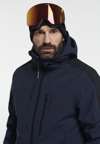 Man in een navy blauwe Tenson skijas, zwarte beanie en reflecterende skibril met een oranje-roze tint, recht vooruit kijkend tegen een witte achtergrond.