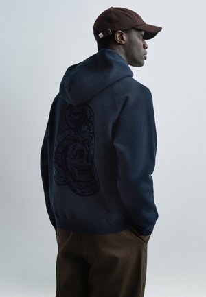 Man draagt een marineblauwe hoodie met een zwart slang- en schedelontwerp op de achterkant, een bruine pet en een bruine broek, kijkt naar rechts met handen in de zakken.