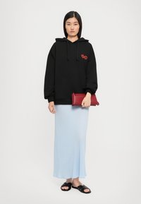 Jovem mulher vestindo um moletom preto com texto vermelho, saia longa azul clara, sandálias deslizantes pretas, segurando uma bolsa de mão vermelha, de pé contra um fundo branco.