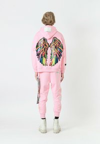 Sudadera rosa con diseño de alas de lentejuelas en la espalda, combinada con pantalones de chándal rosa a juego que presentan acentos bordados y calzado negro.