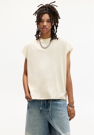 Haut sans manches beige à coupe décontractée, associé à un short en denim bleu clair présentant un léger dégradé et des détails de couture visibles.