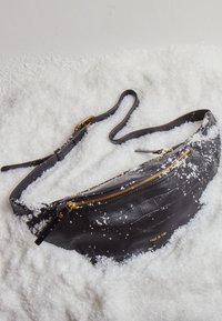 Musta nahkainen vyölaukku krokotiilinkuosilla, kullattu vetoketju, säädettävä hihna, esillä valkoisen lumimaisen materiaalin päällä.