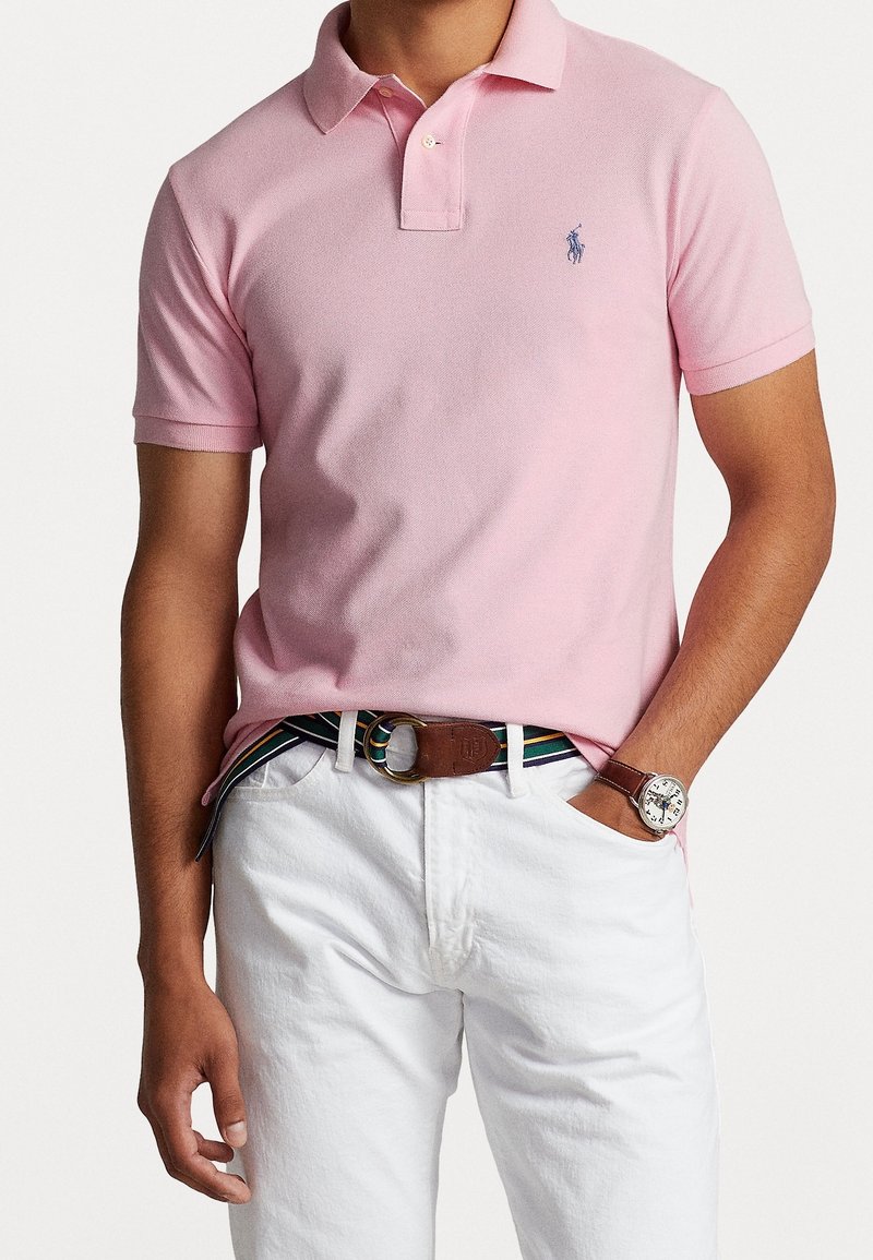 Ljusrosa pikétröja med klassisk krage och korta ärmar, med en liten blå logga. Tillsammans med vita byxor och ett randigt bälte.