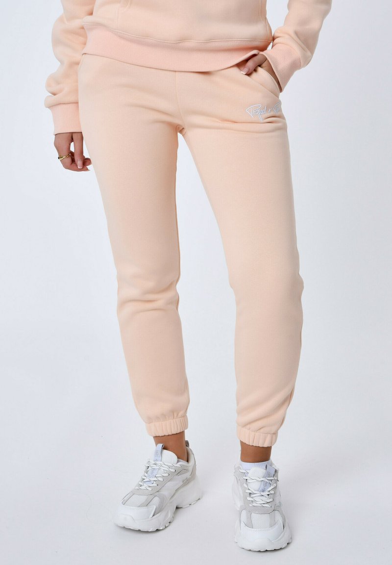 Perzikkleurige sweatpants met een gladde textuur, elastische manchetten en een klein wit logo op de dij, gecombineerd met witte sportschoenen.