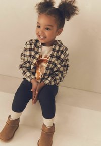 Vaikas vilkintis languotą marškinėlį, baltą marškinį su piešiniu, juodas leginsus, kremines kojines ir rudus aulinukus, sėdi ant baltos paviršiaus.