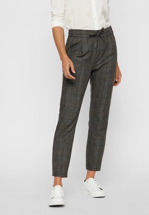 Frau trägt graue, karierte, schmal zulaufende Hose mit Kordelzugbund, weißes Hemd mit Knöpfen und weiße Turnschuhe vor einfarbigem Hintergrund.