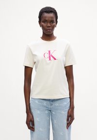 Krems krāsas kokvilnas T-krekls ar īsām piedurknēm, ar lielu rozā Calvin Klein logos centrā. Parādīts ar gaiši ziliem džinsiem.