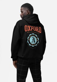 Černá mikina s kapucí s nápisem "OXFORD UNIVERSITY" v tučném oranžovém písmu. Obsahuje dekorativní modrou štítovou logo s písmenem "A" a atletické detaily.