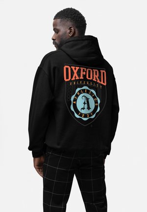 Černá mikina s kapucí s nápisem "OXFORD UNIVERSITY" v tučném oranžovém písmu. Obsahuje dekorativní modrou štítovou logo s písmenem "A" a atletické detaily.