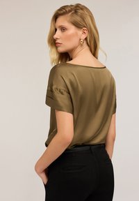 Blusa di raso verde oliva con maniche arrotolate e scollatura ampia, abbinata a pantaloni neri aderenti. Trama liscia, design minimalista.