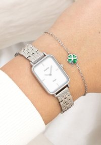 Montre-bracelet en métal argenté avec un cadran blanc rectangulaire et un bracelet en maille, accompagnée d'un bracelet argenté avec un petit pendentif en forme de trèfle à quatre feuilles vert.