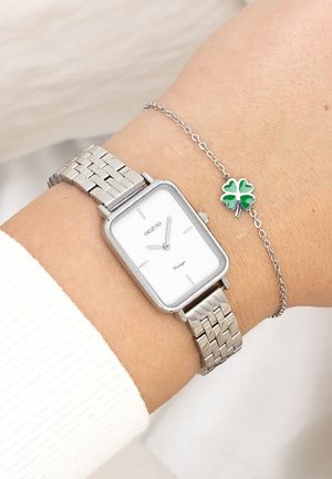 Montre-bracelet en métal argenté avec un cadran blanc rectangulaire et un bracelet en maille, accompagnée d'un bracelet argenté avec un petit pendentif en forme de trèfle à quatre feuilles vert.