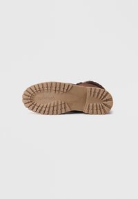 Suela de bota marrón con un diseño de goma texturizada y patrones de suela prominentes, etiquetada con el logo de la marca sobre una superficie de color beige claro.