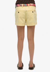 Pantaloni corti di cotone gialli con orli arrotolati, due tasche posteriori con chiusura a bottone e una cintura a fantasia multicolore. Vestibilità piatta e casual.