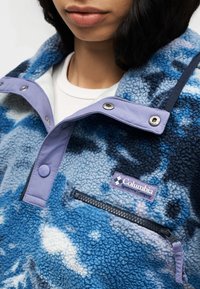 Φλις πουλόβερ σε αποχρώσεις του μπλε με σχέδιο υφής. Διαθέτει γιακά με μωβ κουμπιά τύπου snap, ταμπελάκι με λογότυπο και πλαϊνή τσέπη με φερμουάρ.