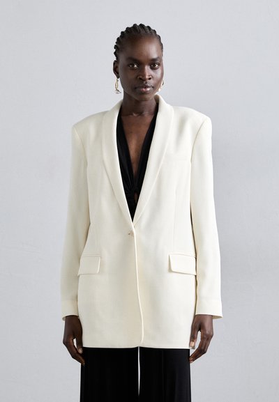 Weißer strukturierter Blazer mit einem einzelnen Knopf, Fronttaschen und einem texturierten Stoff, über einem schwarzen Top getragen von einem Modell.