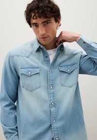 Camicia in denim blu chiaro con due tasche sul petto con chiusura a scatto, colletto classico e maniche lunghe con polsini abbottonati.