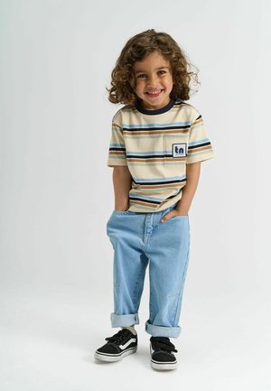 Naeratav laps lokkis juustega seisab käed taskus, seljas triibuline beež särk, helesinised üles keeratud teksapüksid ja mustad spordijalatsid valgel taustal.