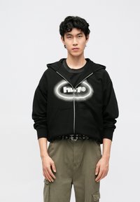 Čierna mikina na zips s predným vačkom typu klokanka a grafickým logom. Nosená cez čierne tričko, skombinovaná s olivovými cargo nohavicami a opaskom.