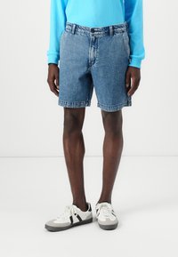 Denim shorts in light blue with a classic cut, featuring a button closure, belt loops, and frayed hems. Paired with white and gray sneakers.
