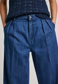 Žena v volných tmavě modrých plisé džínách a námořnickém pleteném topu bez rukávů, se založenýma rukama v kapsách.