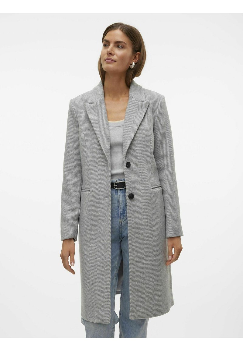 Cappotto grigio a spina di pesce con revers a punta, due bottoni neri e tasche laterali, abbinato a un top grigio e jeans in denim blu.