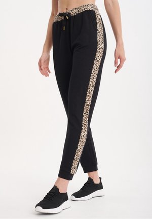 Pantaloni da jogging neri con elastico in vita a stampa leopardata e strisce laterali, indossati con scarpe da ginnastica nere su una persona in piedi.