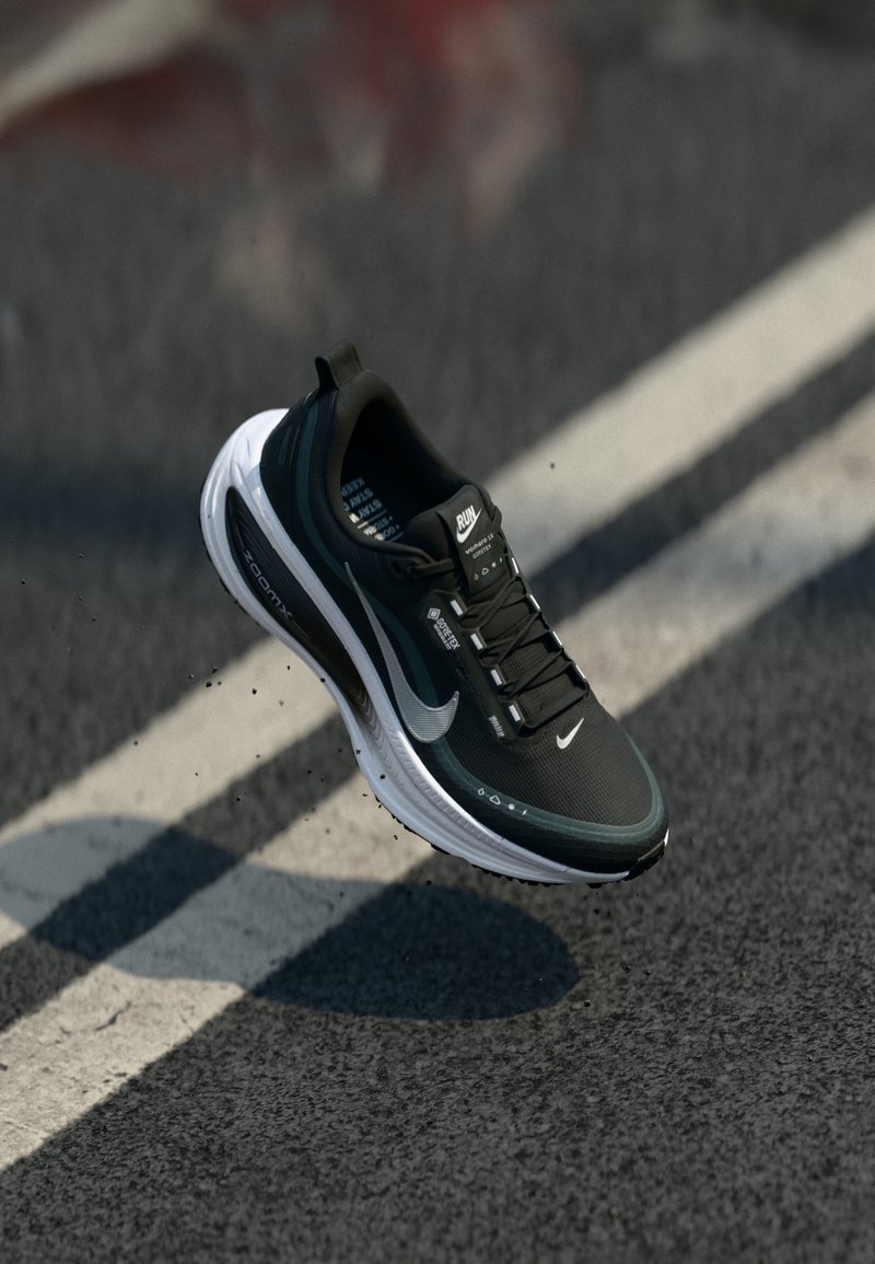 Zwart-witte Nike hardloopschoen in de lucht boven een asfaltweg met een witte streep, met zicht op de Gore-Tex branding en details van de ZoomX zool.