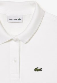 Valkoinen polo-paita, joka on valmistettu teksturoidusta kankaasta ja jossa on kauluspanta, kolme nappia edessä ja vihreä brodeerattu krokotiililogo.