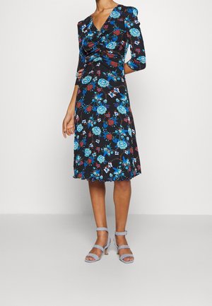 Femme portant une robe noire longueur genou avec imprimé floral bleu et rouge et détail froncé, associée à des sandales à talons hauts bleu clair à brides.