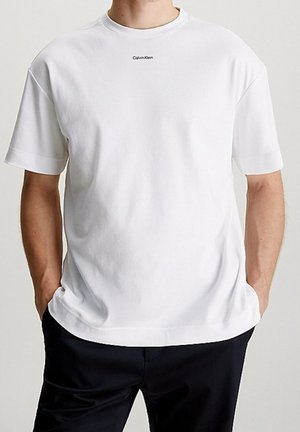 Mężczyzna ubrany w prostą białą koszulkę Calvin Klein z krótkim rękawem, ręce w kieszeniach, w czarnych spodniach na neutralnym tle.